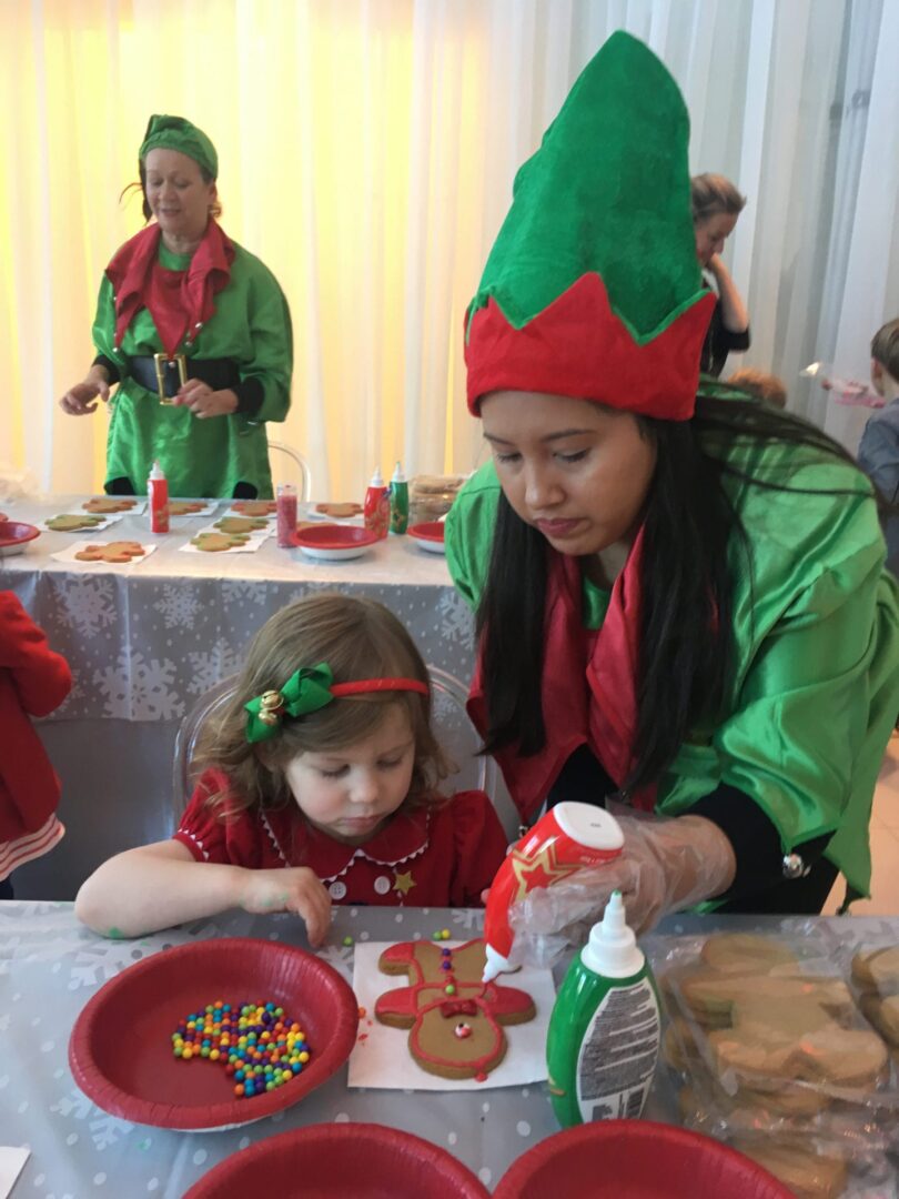 a woman dressed in an elf costume helping a girl decorate a gingerbread cookie
